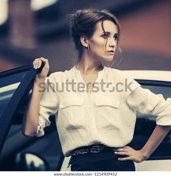 Young fashion business woman next to her car \
Stylish female model with bun up do hair wearing white shirt and\
black pencil skirt
