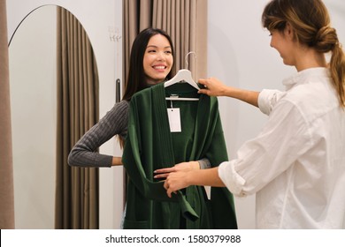 Young Fashion Consultant Helping Trying On Cardigan To Beautiful Smiling Asian Girl In Modern Store