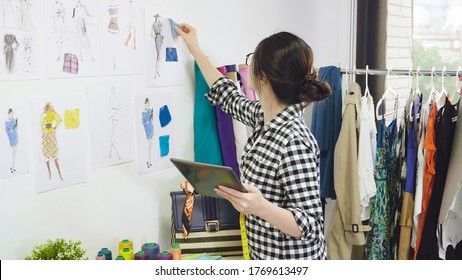 Young Fashion Designer Asian Japanese Woman Looking At Her Tablet Thinking Details Creating In Studio. Dressmaker Hold Digital Touchpad Choosing Color Of Fabric Textile Cloth Sample On Wall Sketch