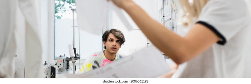 young fashion designer looking at blurred colleague in tailor shop, banner