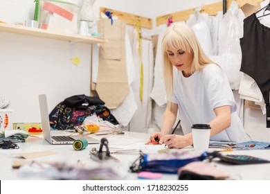 Junge Modedesignerin, die an ihrem Atelier arbeitet. Fashion Design Studio mit arbeitendem Personal Computer, Hanging Kleidung, Kaffee und verschiedene Nähartikel auf dem Tisch. Frau kreiert stylische Kleidung