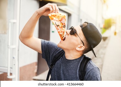 Young fashion fun hipster guy eating pizza outdoors. Sunglasses, cap, on the hand watch, a great carefree day, urban style