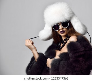 Young fashion girl in fur coat and white hat in modern round sunglasses on dark background