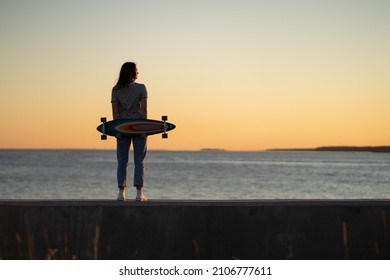 Young fashion girl with longboard enjoy sunset looking at seaside skyline. Stylish trendy female longboarder silhouette over beautiful setting sun at sea coast. Freedom, youth and lifestyle concept