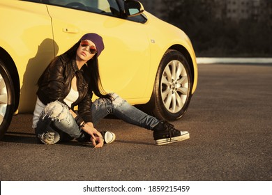 Young fashion hipster woman sitting on sidewalk leaning on car  Stylish female model in black leather jacket purple beanie and ripped jeans 