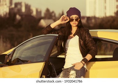 Young fashion hipster woman in sunglasses leaning on her car  Stylish female model in black leather jacket purple beanie and ripped jeans 