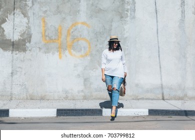 Young fashion woman wearing ripped jeans, colorful heels and straw accessories posing over gray concrete city wall. Plus size model.