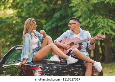 Young fashionable couple playing acoustic guitar on the back side of their convertible car in nature. - Powered by Shutterstock