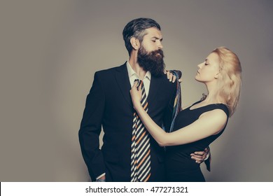 Young Fashionable Couple Of Woman With Pretty Face And Blonde Hair And Handsome Bearded Man With Long Beard In Black Jacket And Tie In Studio On Grey Background