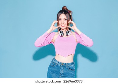 Young fashionable cute carefree Asian woman wearing headphone and roller skate with perfect slim body showing heart sign on isolated blue background. Female model posing indoors studio.