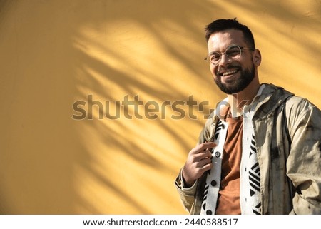 Young fashionable hipster man posing outdoor with yellow wall background. Style and independence millennial male standing for portrait. World traveler youth culture