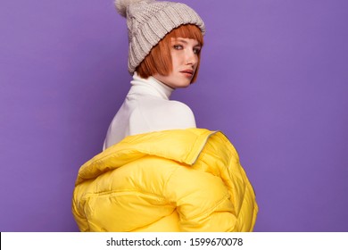 Young Fashionable Red Hair Woman Wearing Nice Yellow Down Jacket, Warm Winter Hat. Portrait Of Happy Smiling Girl In Stylish Glamorous Clothes. Autumn Winter Fashion Photo On A Purple Background.