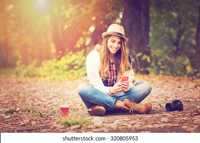 Young fashionable teenage girl with smartphone, camera and takeaway coffee in park in autumn sitting at smiling. Trendy young woman in fall in park texting. Retouched, vibrant colors. - Powered by Shutterstock