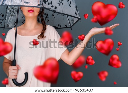 Young fashionable woman holding umbrella standing against grey background red hearts are floating around her. Love rain on Saint Valentine`s Day concept