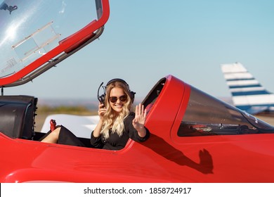 Young Fashionable Woman Pilot In Headset Ready To Fly In Small Red Airplane. Beautiful Life, Aristocratic Lady In Black Dress In Blonde Wavy Hair.