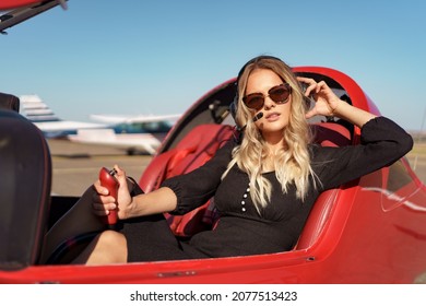 Young Fashionable Woman Pilot In Headset Ready To Fly In Small Red Airplane. Beautiful Life, Aristocratic Lady In Black Dress In Blonde Wavy Hair.