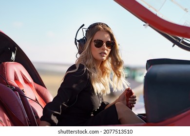 Young Fashionable Woman Pilot In Headset Ready To Fly In Small Red Airplane. Beautiful Life, Aristocratic Lady In Black Dress In Blonde Wavy Hair.