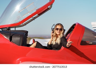 Young Fashionable Woman Pilot In Headset Ready To Fly In Small Red Airplane. Beautiful Life, Aristocratic Lady In Black Dress In Blonde Wavy Hair.