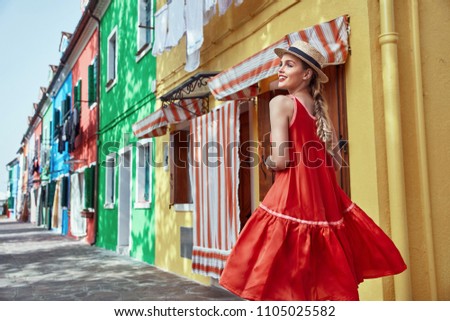  Young fashionable woman in romantic red dress and straw hat, with braid hairstyle, posing near colorful houses in Burano Island, Venice, Italy  
