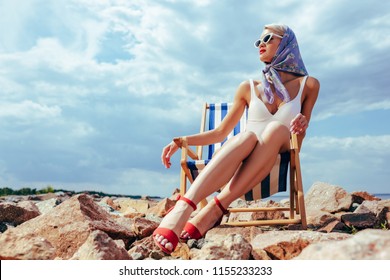 Young Fashionable Woman In Vintage Swimsuit Resting In Beach Chair On Rocky Shore