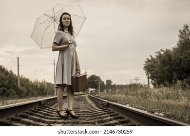 A Young Fashionista Woman In Vintage Style With A Suitcase And A Summer Umbrella Stands On The Railway And Looks Ahead Purposefully.