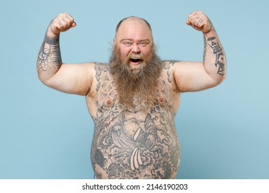 Young fat pudge obese chubby overweight blue-eyed bearded man 30s has big belly with naked tattooed torso showing biceps muscles on hand demonstrating strength power isolated on pastel blue background