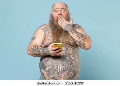 Young fat pudge obese chubby overweight blue-eyed bearded man 30s has big belly with naked tattooed torso hold mobile cell phone covering mouth with hands isolated on pastel blue background studio.