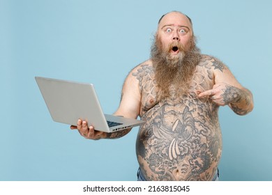 Young Fat Pudge Obese Chubby Overweight Blue-eyed Bearded Man 30s Has Big Belly With Naked Tattooed Torso Point Finger On Laptop Pc Computer Chatting Online Isolated On Pastel Blue Background Studio.