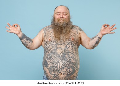 Young fat pudge obese chubby overweight blue-eyed bearded man 30s has big belly with naked tattooed torso hold hands in yoga om gesture relax meditate try calm down isolated on pastel blue background.