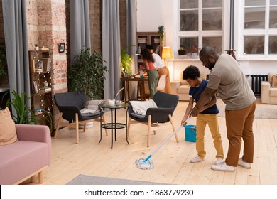 Young Father Of African Ethnicity Showing His Little Son How To Wash The Floor In Living-room While Mother And Daughter Cleaning Table Together