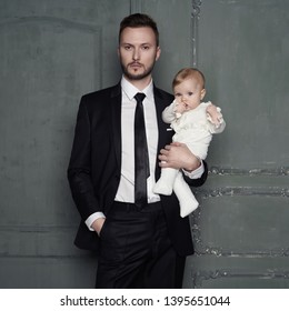 Young Father With Beautiful Little Baby In His Arms. Fashion Portrait Of Man With Pretty Daughter. Stylish Dad In Evening Black Suit And Small Girl In White Sliders. Studio Portrait.