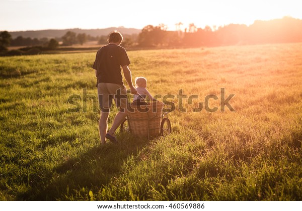 Young father is\
carring his son in the garden cart in the green meadow with the\
sunset. Young man and the child enjoying summer evening outdoors.\
Sunshine. Color toned\
image.