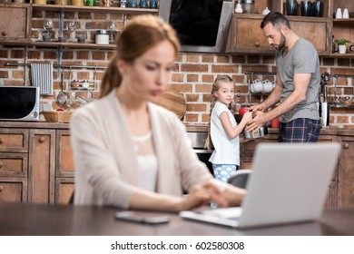 Young Father And Child Girk Making Tea While Woman Working On Laptop