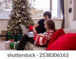 Young father and daughter in plaid pajamas enjoy heartwarming moment on sofa, with laptop, wrapped red gift, surrounded by holiday cheer, decorated Christmas tree. Shopping online, sales