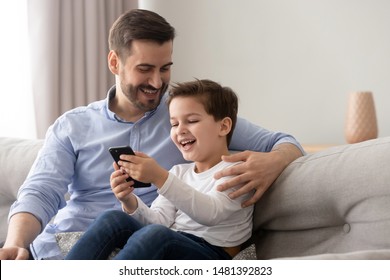 Young father embraces little son family sitting on couch at home using smart phone feels happy cheerful laughing on pranks online funny videos, parental control and modern tech everyday usage concept - Powered by Shutterstock