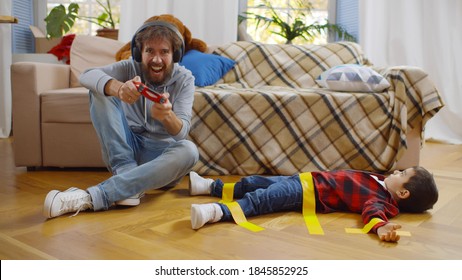 Young Father In Headphones Playing Video Game With Console On Disobedient Son Tied With Duct Tape On Floor. Dad Looking After Kid Alone In Living Room