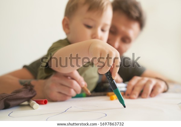 Young father helps son at age of two draw, teaches\
alphabet. A small boy is sitting on his dads lap near white wall in\
room at table, on piece of paper writing letters and pictures with\
felt-tip pen.