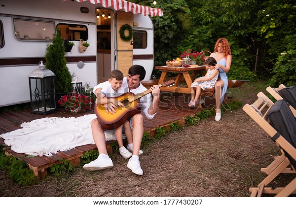 A young father with his little son play the\
guitar together in front of their trailer house on the background\
at a wooden table a beautiful mother with red hair sits, weaves\
braids to tender daughter