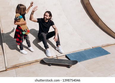 The young father and his son dressed in the stylish casual clothes are sitting and have fun together on the slide next to the skateboards in a skate park at the sunny warm day - Powered by Shutterstock