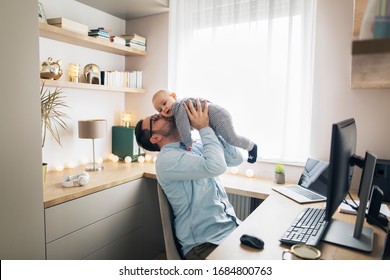 Young father holds his baby boy and kisses him while sitting in front of computer trying to work from home. - Shutterstock ID 1684800763