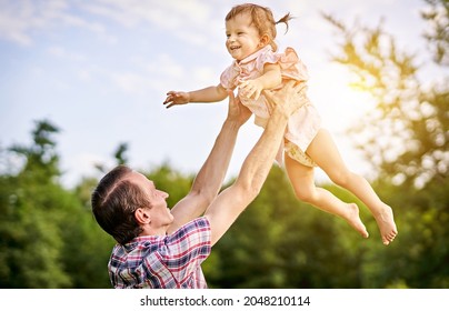 Young Father Lifting Up Her Toddler Laughing Little Daughter Overhead In The Air - Dad And His Baby Playing Together Outdoors In Sunset - Paternity And Childhood Concept