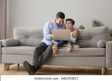 Young father and little son sitting on couch in modern cozy living room spend free time at home having fun using notebook laughing watching comedy funny movie, computer games online amusements concept