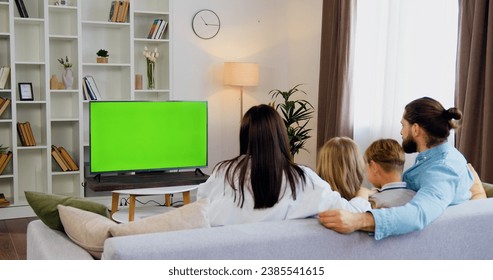 Young father and mother son and daughter sit on the couch and watch green screen TV with fun with smiles. Beautiful family is watching TV on chroma key at home in living room.