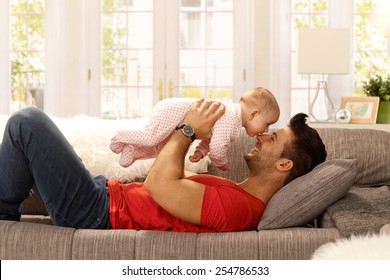 Young Father Playing With Baby Daughter As Lying On Sofa At Home, Smiling Happy Having Fun. Side View.