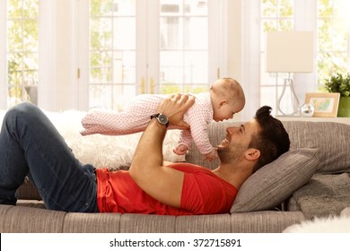 Young Father Playing With Baby Daughter On Sofa. Side View.