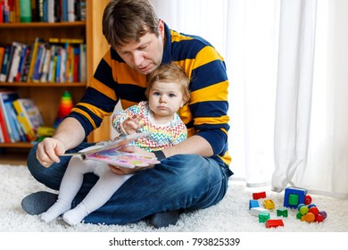 Young Father Reading Book With His Cute Adorable Baby Daughter Girl. Smiling Beautiful Child And Man Sitting Together In Living Room At Home. Toddler Hearing To Dad