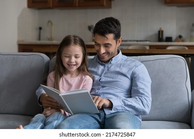 Young father reads a book to little 6s cute daughter sit together on sofa spend leisure enjoy good life hobby and pastime at home. Fatherhood, upbringing, children development, intelligence concept - Shutterstock ID 2174725847
