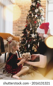 Young Father In Red Santa Hat Giving Christmas Gift To His Smiling Daughter Near Decorated Christmas Tree. Girl Dressed In Festive Red-black Christmas Outfit. They Smiling, Happy Because Of New Year