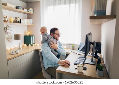 Young father sitting in front of computer trying to work from home while holding his baby boy.