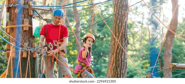 Young father teaching her daughter to climb on the rock climbing wall. Little girl preschooler wearing safety harness having fun time in adventure rope park. Happy family concept - Powered by Shutterstock